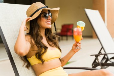 Dışarıda bikinisiyle güneşlenirken şapka ve güneş gözlüğü takarak kokteyl içen mutlu genç bir kadın.