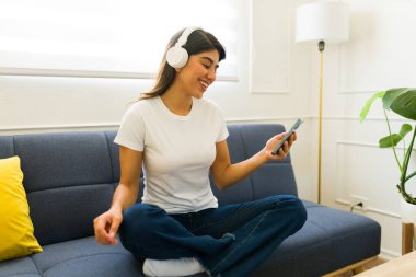Latin genç bir kadın evde kulaklıkla müzik dinlerken beyaz bir tişörtle akıllı telefonunu kullanıyor.