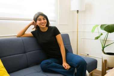 Güzel İspanyol kadın kanepede rahatlarken gülümsüyor. Evinde siyah bir tişört giyiyor.