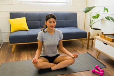 Rahatlamış genç bir kadın evde gri bir tişört giyerek yoga yaparken meditasyon yapıyor ve dinleniyor.