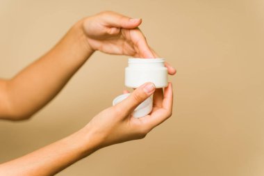 Genç kadın elleri cilt bakım ürünü tutuyor ve stüdyo arka planına krem nemlendirici sürüyor.