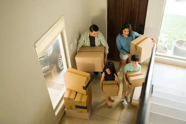 Latin family carrying cardboard boxes with the help of the kids while unpacking and moving into a new beautiful house