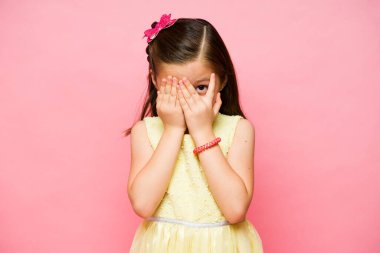 Korkak küçük ilkokul kızı gözlerini kapatıyor. Pembe arka plandan korkan korkutucu bir şeye bakıyor.