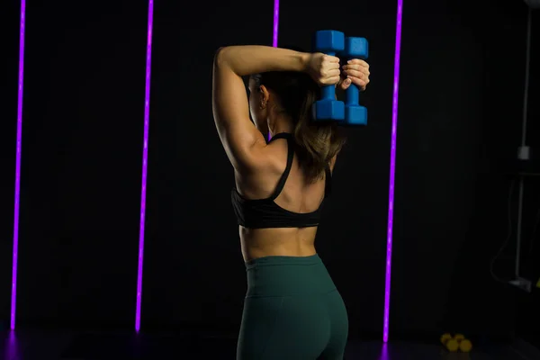 一个适合运动的年轻女子练哑铃举重和在一个黑暗的高端健身中心锻炼的后视镜 — 图库照片