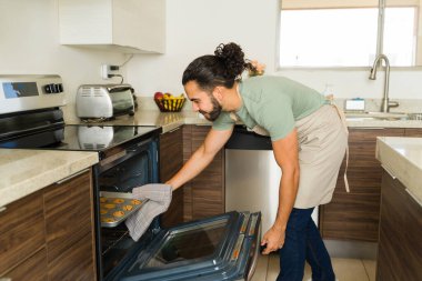 Yakışıklı Meksikalı adam mutfağının elektrikli fırınında kurabiye pişirirken gülümserken ev işi ve ev hayatından zevk alıyor.
