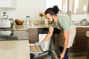 Neşeli gülümseyen bir genç adam elektrikli fırında lezzetli kurabiyeler pişirirken mutfakta ev işlerini yaparken mutlu görünüyor.