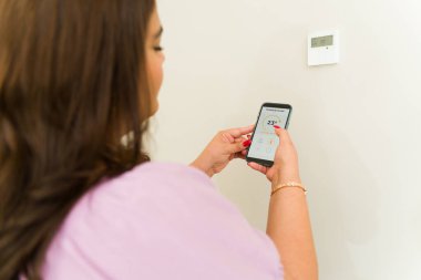 Evdeki akıllı telefon uygulamasını kullanırken termostatı ve klimayı kontrol eden genç bir kadının arka görüntüsü