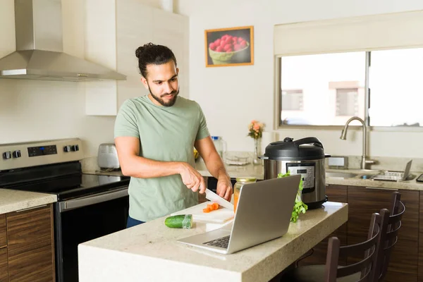Handsome mexican man cutting vegetables in the kitchen and cooking food for lunch while watching a video tutorial on the laptop following a recipe