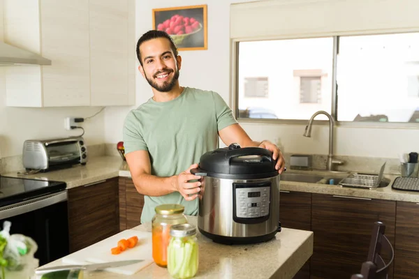 Cheerful hispanic man smiling in the kitchen happy to cook dinner using the pressure cooker or slow cooking pot in his beautiful kitchen