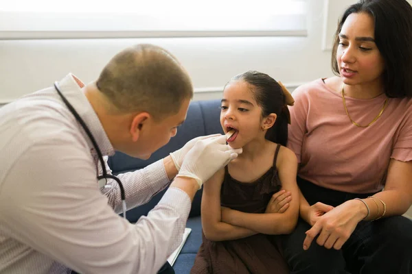 Pediatrician using a tongue depressor checking the sore throat of a sick child during a medical visit at home