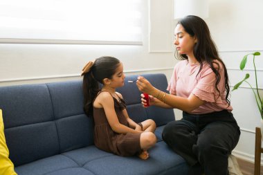 Evde yorgun Latin bir anne hasta çocuğuna öksürük şurubu ve ilaç veriyor ve kızına bakıyor.