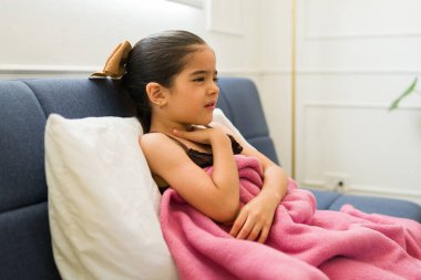 Soğuk algınlığı ya da evde dinlenen bir battaniyeye sarılmış grip yüzünden boğaz ağrısı çeken hasta bir çocuk.