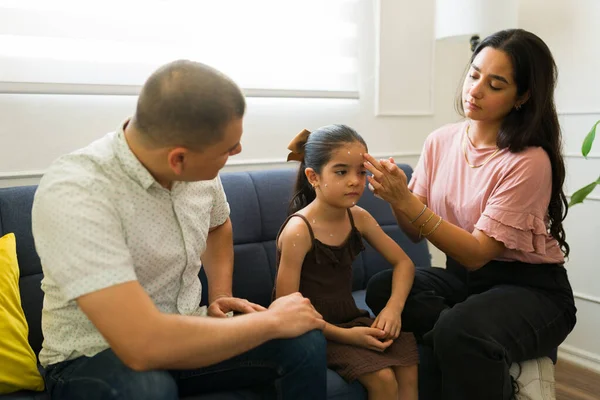 Latin parents looking worried while putting ointment cream on the face of their young daughter sick with chickenpox