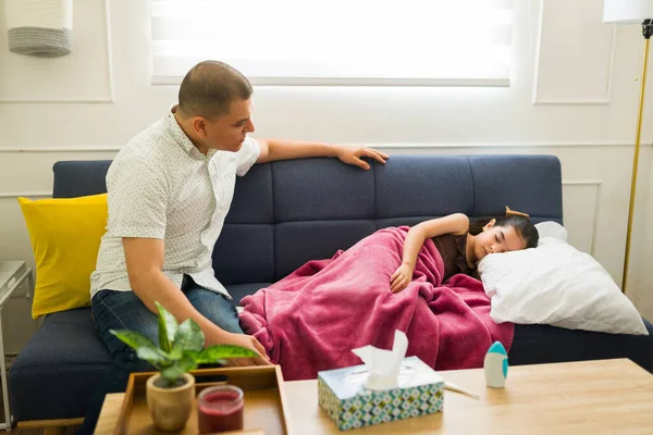 Latin father checking on her sick little daughter sleeping on the couch with a blanket resting from a fever or flu