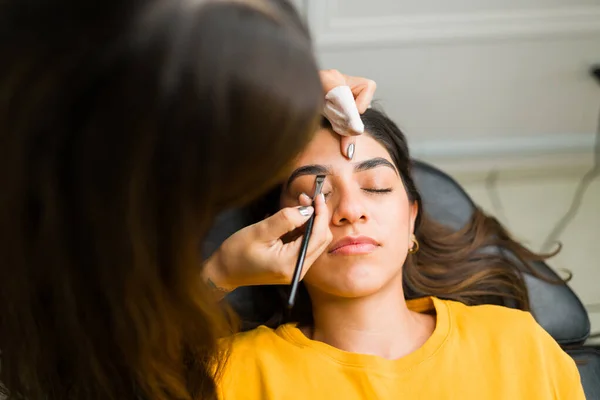 Attractive woman customer relaxing while in the beauty salon with the cosmetologist doing a new brow design with henna