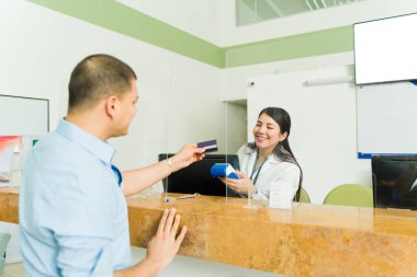 Tıbbi laboratuvarda çalışan neşeli bir kadın kan testleri için kredi kartıyla ödeme yapan bir hastayla çalışıyor.