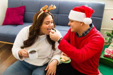 Romantik latin adam büyük beden kız arkadaşına lezzetli kurabiyeler veriyor ve Noel 'de oturma odasında sıcak çikolata içiyor.
