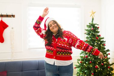 Heyecanlı ve kilolu bir kadın çirkin bir kazakla dans edip Noel ağacının önünde tatil müziği dinlerken eğleniyor.