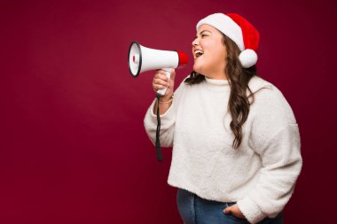 Noel Baba şapkalı güzel obez bir kadının profili. Bir megafon kullanarak yılbaşı mesajı bağırıyor. Heyecanlı görünüyor.