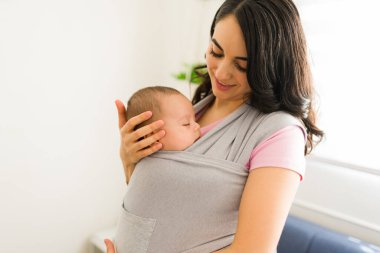 Gülümseyen mutlu anne, evde bebek askısı taşırken bebeğini çok seviyor.