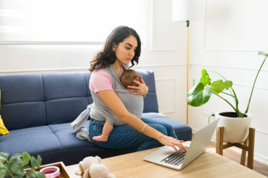 Profesyonel bir anne oturma odasında oturmuş yeni doğmuş bebeğini kucağında bebek askısıyla uzaktan kumandayı kullanıyor.