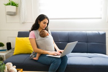 Çalışan anne dizüstü bilgisayarda yazarken gülümsüyor. Evde uzaktan çalışıyor. Çocuğuna bebek askısıyla bakıyor.