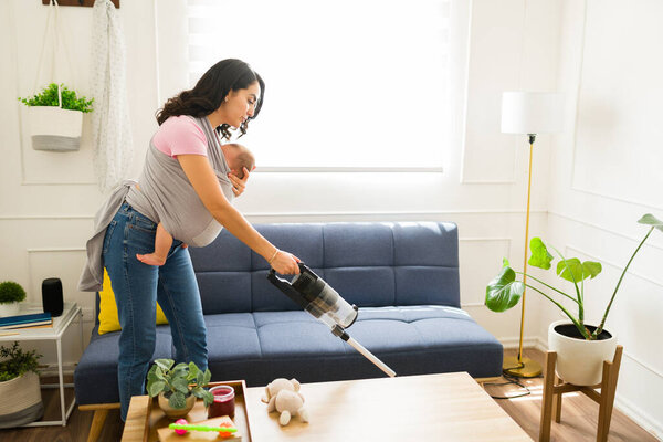 Кавказская мама убирает дом и делает домашнюю работу, пылесошая гостиную и вынашивая ребенка с помощью детской повязки