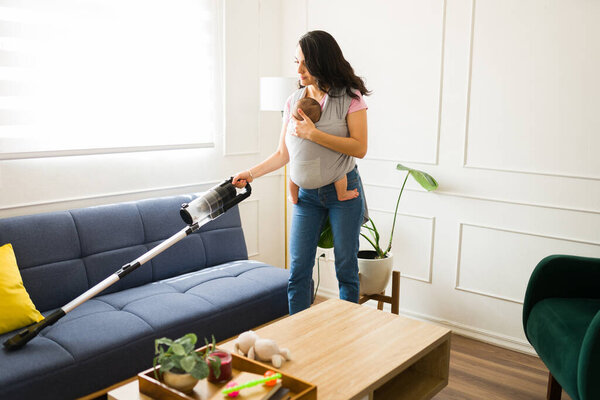Мать носит своего новорожденного ребенка с помощью детской стропы пылесосить гостиную и делать уборку по дому