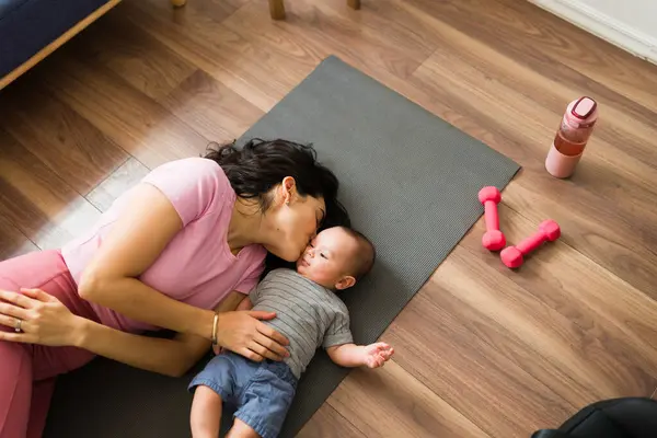 Relaxed loving mom kissing her adorable newborn baby resting on the floor after exercising and doing a home workout