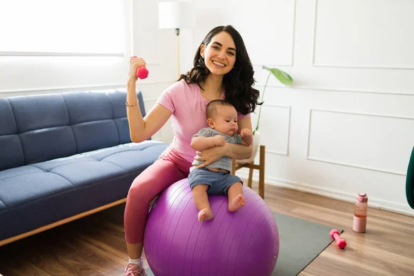 ダンベルウェイトと安定性ボールで働く自宅で彼女の新生児と陽気で美しい母親 — ストック写真