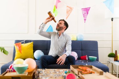 Oturma odasındaki dağınık evde alkollü içki içen, boş bira şişesi olan sarhoş beyaz bir adam.