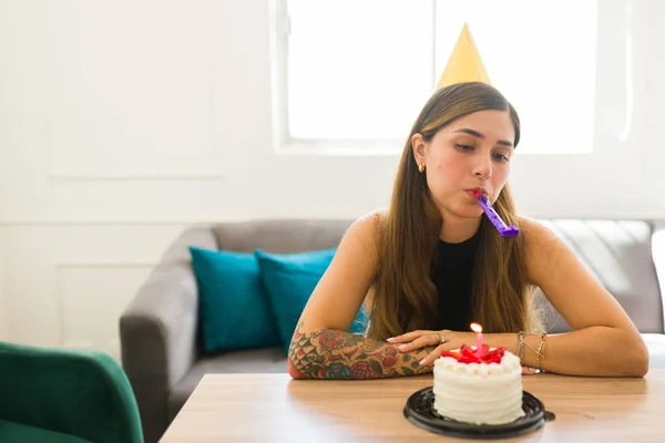 Depressed latin woman feeling sad while using a party whistler to celebrate her birthday alone at home and eating cake