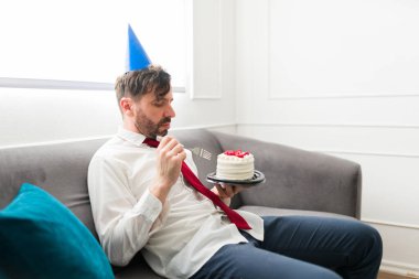 Üzgün bunalımlı adam kanepede tek başına oturmuş tek başına doğum gününü kutluyor ve doğum gününü parti şapkasıyla yiyor.