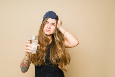 Hasta Meksikalı kadın baş ağrısı çekiyor ve sabahları akşamdan kalma görünüyor. Uyku maskesi takıp ilaçla su içiyor.