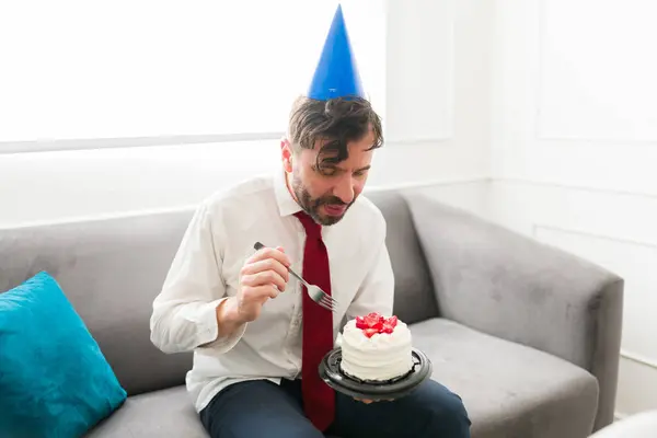 Attractive caucasian man eating cake alone wearing a party hat celebrating his birthday alone looking sad