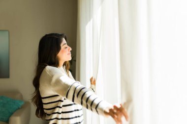 Evde güneşli ve rahat bir sabahta gülümseyen ve rahatlatan Latin bir genç kadın pencereleri açıyor.