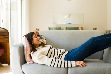 Gülümseyen mutlu genç kadın kanepede uzanıp rahat evinde dinlenirken sağlıklı yaşam tarzının tadını çıkarıyor.