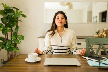 Rahatlatıcı bir meditasyon yapan ve evde çalışırken stressiz bir hayat ve sağlıklı bir yaşam süren rahat Latin bir genç kadın.
