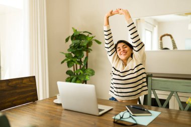 Mutlu, rahat, genç bir kadın dizüstü bilgisayarda çalışırken sırtını ve kollarını uzatıyor. Uzaktan çalışıyor ve evde gülümsüyor.