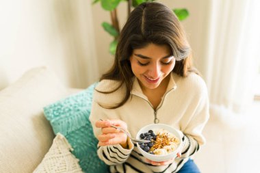 Sağlıklı bir yaşam tarzı yaşarken evde meyve dolu sağlıklı bir yoğurt kasesi yerken gülümseyen İspanyol genç bir kadının en iyi görüntüsü.