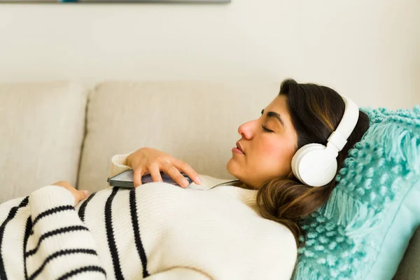 Profile of a latin relaxed young woman listening to a guided meditation with headphones practicing mindfulness while relaxing on the couch