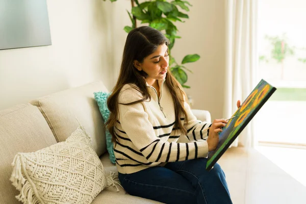 Relaxed hispanic young woman with a wellness lifestyle relaxing on the sofa and painting enjoying her art hobby in her cozy home