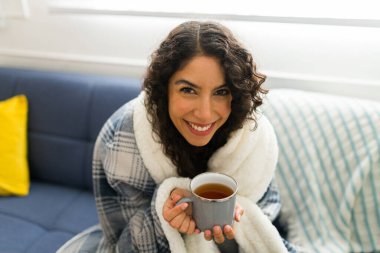 Güzel Latin bir kadın kışın dinlenirken battaniyeye sarılı papatya çayı içerken göz teması kurarak gülümsüyor.