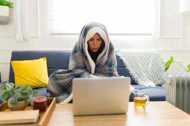 Sıcak bir battaniyeye sarılı üzgün genç bir kadın. Dizüstü bilgisayar kullanırken ya da kış havasında evde online alışveriş yaparken çok üşüyor.