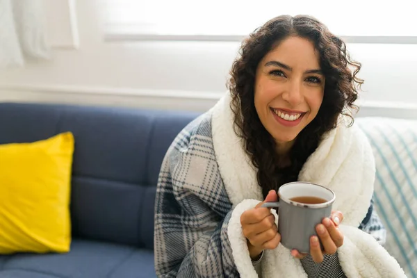 Relaxed happy young woman wrapped in a warm blanket while relaxing on the sofa at home during winter weather eating hot chocolate or tea