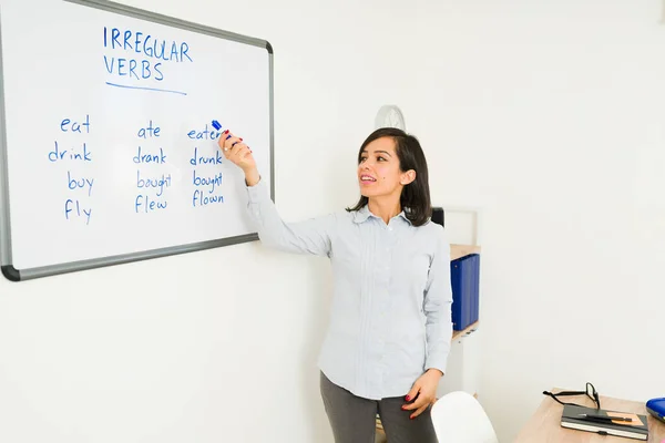 Smart female teacher teaching a class lesson writing on the whiteboard teaching about English irregular verbs