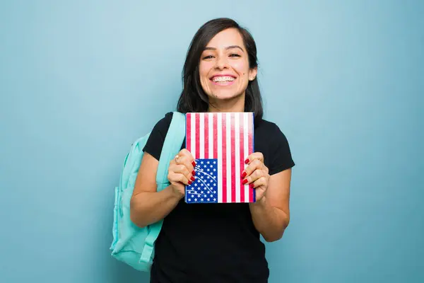 Attractive woman student learning English carrying a backpack holding a notebook with the US flag in front of the studio blue background