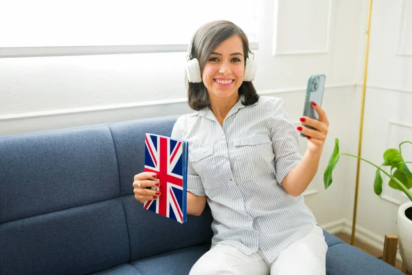 Gorgeous woman with headphones using a book with the UK flag learning english online using a learning app on the smartphone