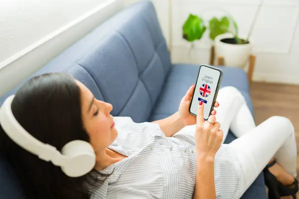 Woman in her 30s relaxing on the sofa with headphones using the online app on her smartphone to learn English in the UK
