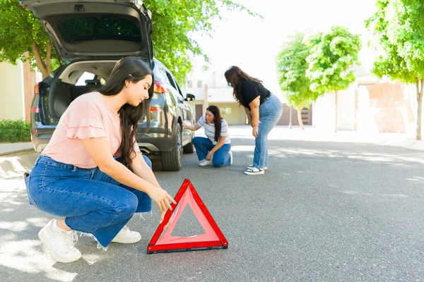 美しい若い女性は 彼女の友人と運転しながら彼女の車の平らなタイヤを変更する道路上の三角信号を置く — ストック写真
